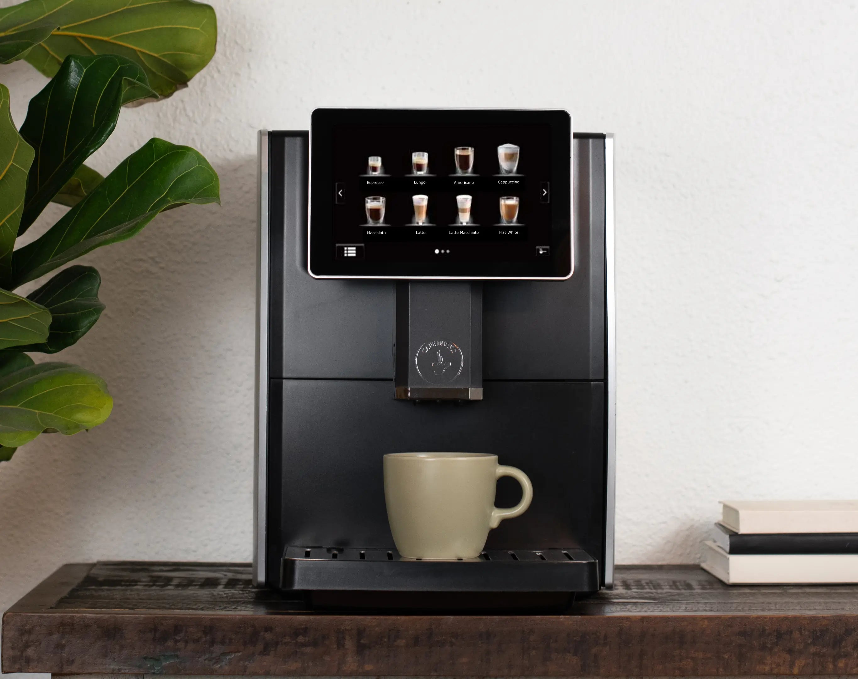 Cafe Bueno CB-3000 super automatic espresso machine front view in matte black and brushed silver on dark wood surface, touchscreen displaying espresso lungo americano cappuccino macchiato latte latte macchiato and flat white drink options, sage green ceramic mug on drip tray, fiddle leaf fig plant on left, stacked books on right, white textured wall background.