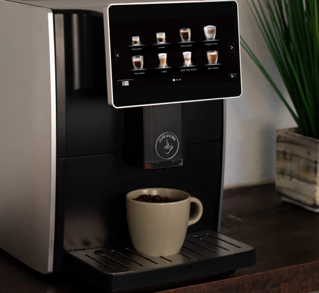 Cafe Bueno super automatic espresso machine front view with large touchscreen displaying eight drink options including espresso lungo americano cappuccino macchiato latte latte macchiato and flat white, cream ceramic mug on drip tray, Cafe Bueno logo on dispenser, brushed silver and matte black finish, dark wood surface with potted grass plant in background.