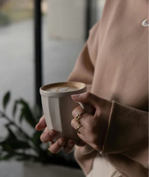 Person in beige sweatshirt holding a white ceramic mug of latte with latte art, wearing gold stacked rings, with a blurred plant and window in the background, warm cozy lifestyle coffee moment.