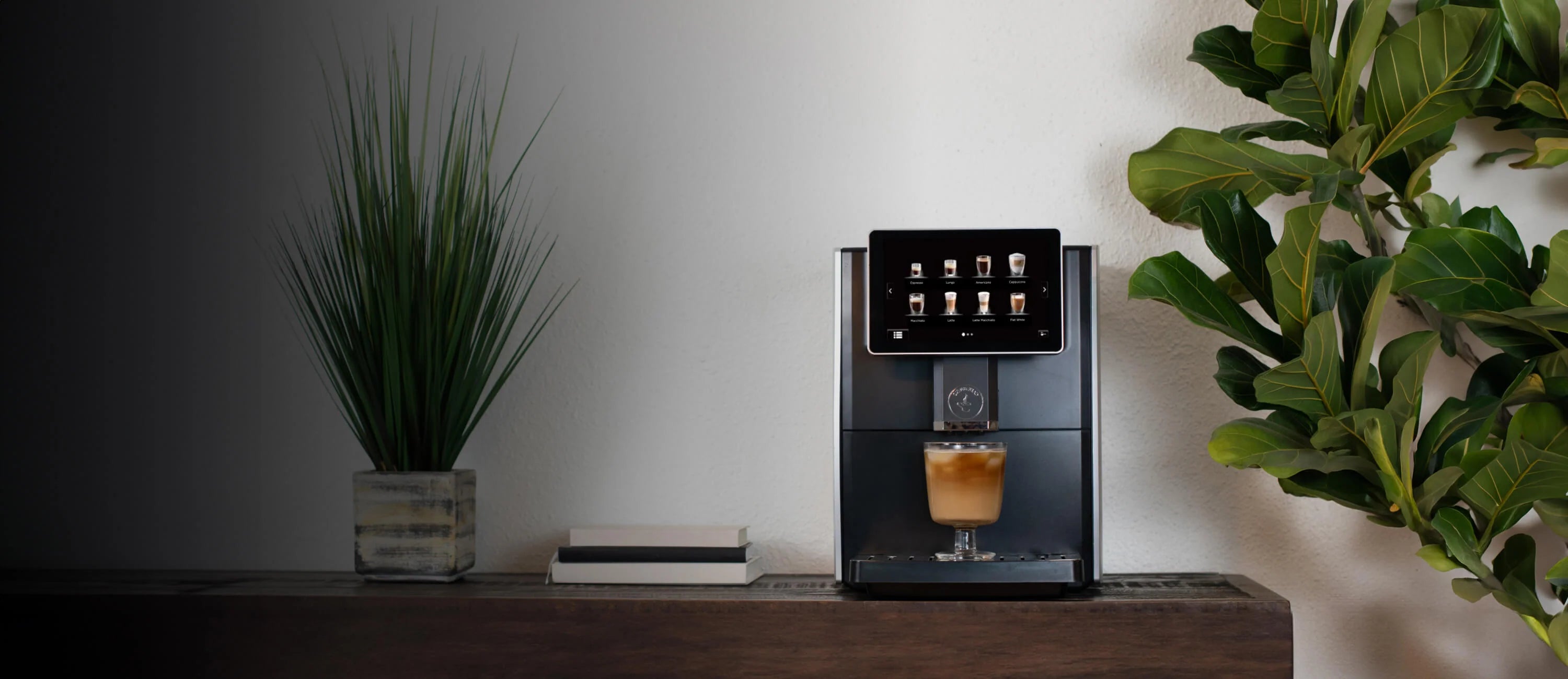 Cafe Bueno CB-3000 bean to cup espresso machine in matte black with silver trim, centered on a dark walnut shelf with an iced latte in the drip tray, flanked by a potted grass plant and stacked books on the left and a fiddle leaf fig on the right, against a white wall with dramatic dark gradient lighting