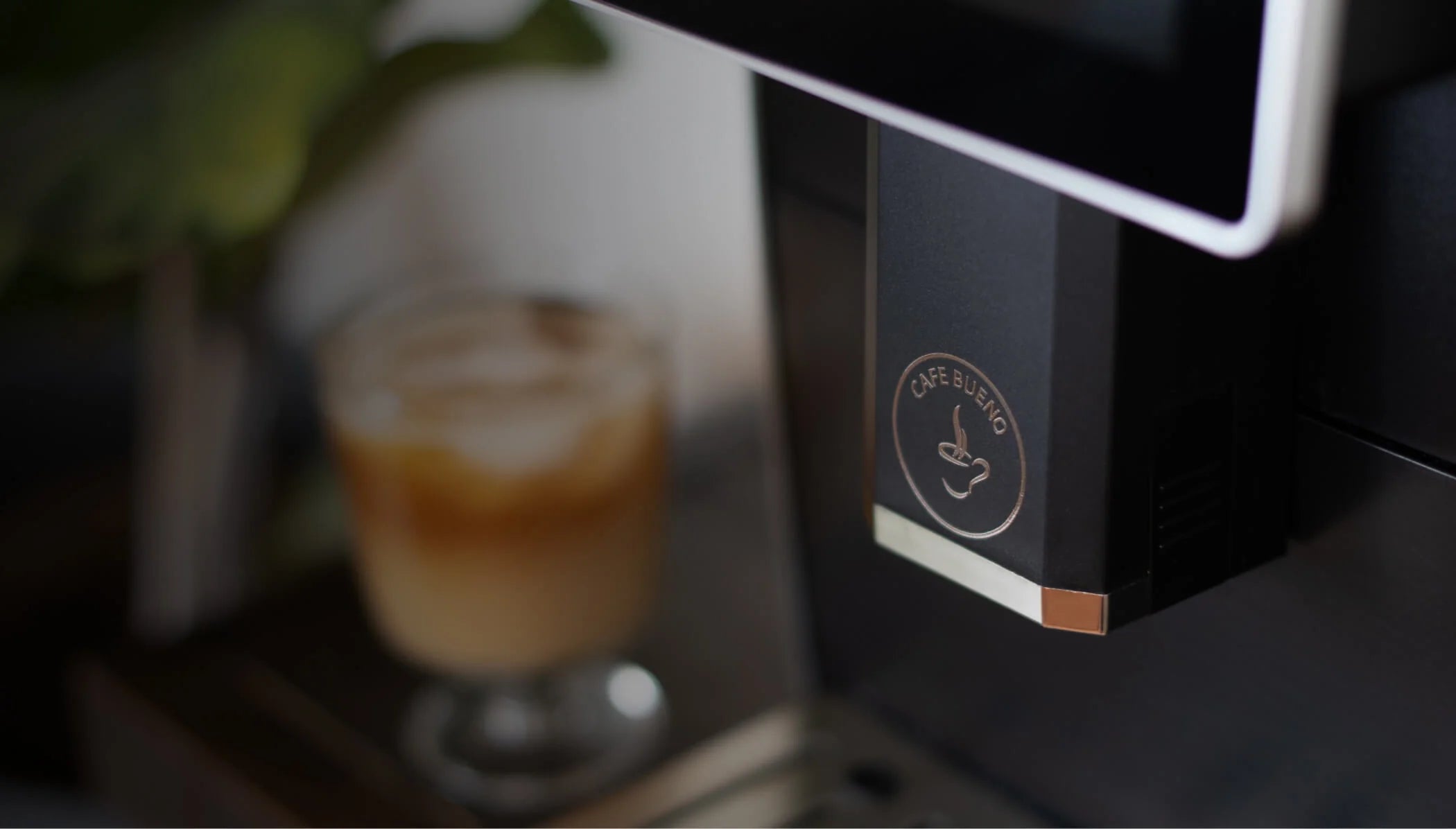 Close-up of Cafe Bueno espresso machine body showing embossed brand logo with coffee cup icon in rose gold on matte black finish, with an iced coffee drink and plant blurred in the background.